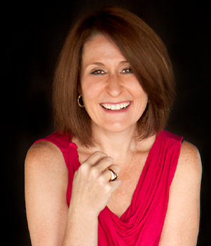 Smiling woman with shoulder-length brown hair in a red sleeveless top against a dark background.