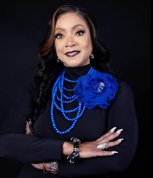 Portrait of a woman with dark hair wearing a black top, layered blue bead necklaces and a blue flower brooch, arms crossed against a dark background.