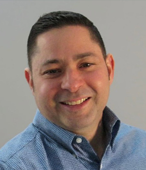 Smiling man with short dark hair in a blue button-up shirt against a gray background.