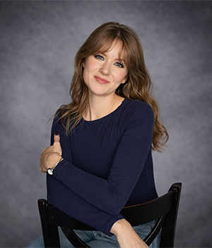 Smiling woman with light brown hair in a navy sweater sits on a black chair against a gray studio backdrop.