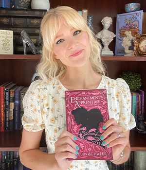 Blonde woman in a floral blouse smiles while holding a pink book titled 'Enchantments Apprentice' in front of a bookshelf.