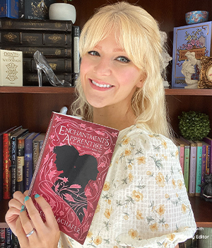 Smiling blonde woman holding a pink copy of 'Enchantments & Apprentices' in a cozy bookstore setting.