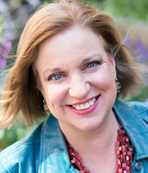Close-up portrait of a smiling woman with blue eyes and short light brown hair, wearing a teal jacket and red beaded necklace outdoors.