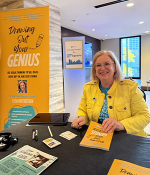 Smiling woman in a yellow jacket sits at a table at a book signing booth, signing copies of the book 'Drawing Out Your Genius' with a bright yellow promotional banner nearby.