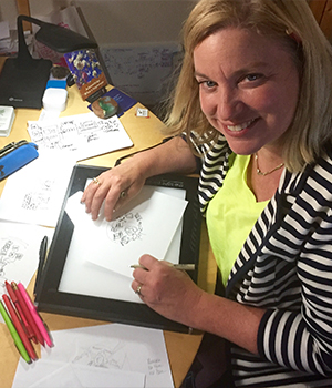 Woman sits at a desk drawing sketches on a lightbox, smiling at the camera while workspace supplies surround her.