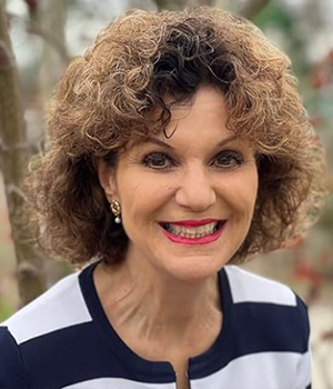 Smiling woman with short curly brown hair and pearl earrings, wearing a navy-and-white striped top, outdoors with greenery in the background.