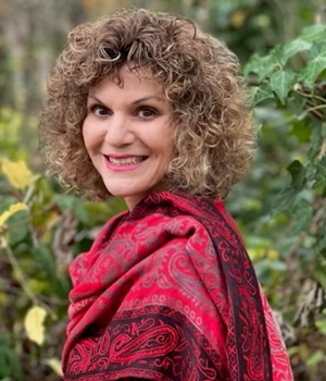 Portrait of a smiling woman with curly hair wearing a red patterned shawl outdoors in a garden, with green plants in the background.