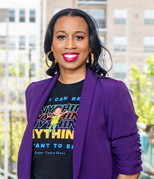 Portrait of a smiling woman wearing a purple blazer, hoop earrings, and a black graphic t-shirt, outdoors in an urban setting.
