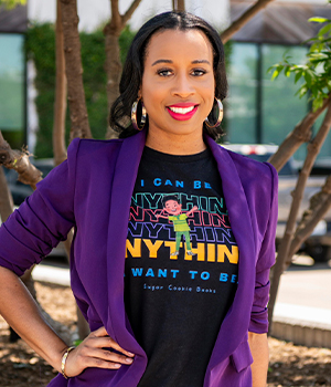 Confident woman in a purple blazer over a black graphic tee with colorful 'ANYTHING' text, posing outdoors with trees in the background.