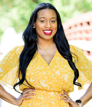 Smiling woman with long dark hair wearing a yellow patterned dress, standing outdoors with hands on hips.