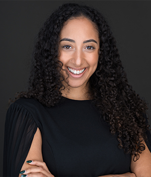 Portrait of a smiling woman with long curly dark hair, wearing a black top, arms crossed, against a dark backdrop.