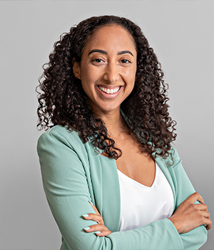 Smiling woman with curly hair in a mint blazer and white top, arms crossed, against a light gray background.