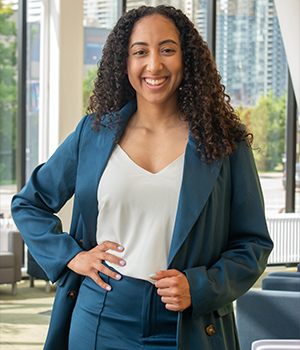 Professional woman with curly hair smiling, wearing a blue blazer and white blouse, standing in a modern office space with large windows.