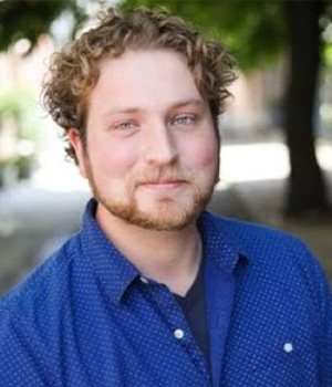 Portrait of a man with curly hair and a beard wearing a blue shirt outdoors and looking at the camera.
