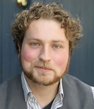 Portrait of a man with curly light brown hair and a short beard, wearing a gray vest and striped shirt, facing the camera.