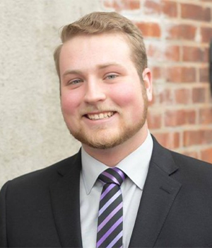 Head-and-shoulders portrait of a smiling man in a dark suit, light shirt, and purple striped tie, standing against a brick wall.