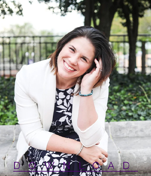Smiling woman in a white blazer sits in a park, resting her head on her hand.