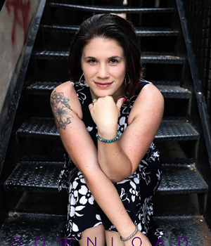 Portrait of a woman sitting on metal stairs, wearing a black-and-white floral dress, with a tattoo on her left arm and bracelets, looking at the camera.