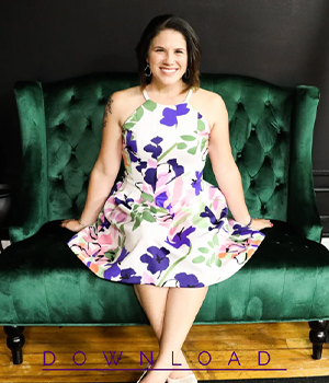 Smiling woman in a white floral dress sits on a green velvet tufted couch, posing for a photo.