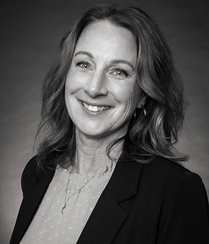 Professional headshot of a smiling woman with shoulder-length wavy hair, wearing a black blazer against a neutral backdrop.