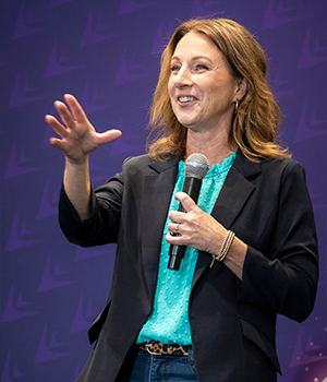 Woman in a teal blouse and dark blazer speaking on stage, holding a microphone and gesturing with her left hand.