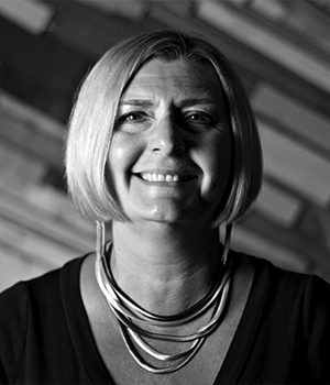 Portrait of a smiling woman with short blonde hair wearing a layered necklace, in black and white.