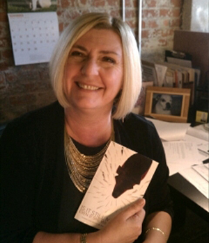 Smiling blonde woman in a black blazer sits at a desk in an office, holding a book with a white cover.