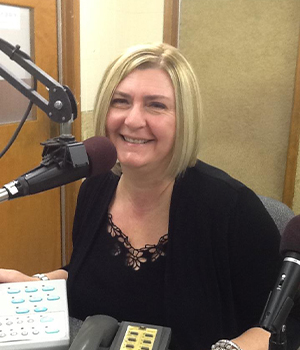 Smiling blonde woman in a black lace top sits at a radio studio desk with a microphone in front.